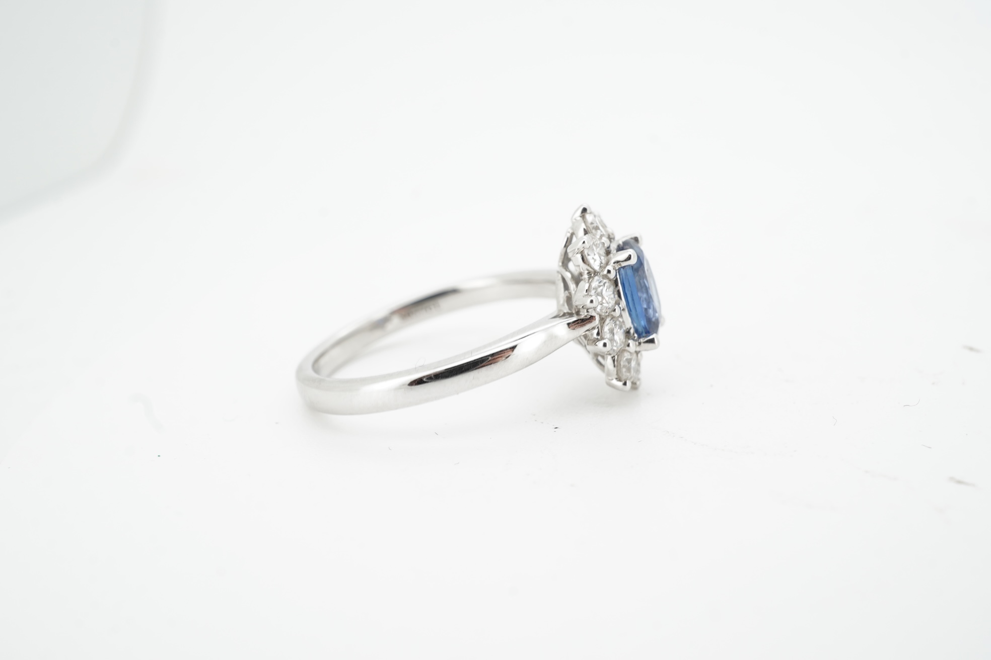 A sapphire and diamond cluster ring, claw-set with an oval sapphire weighing approximately 0.77 carats, within a border of brilliant-cut diamonds totalling approximately 0.50 carats, mounted in white gold, size M, stampe